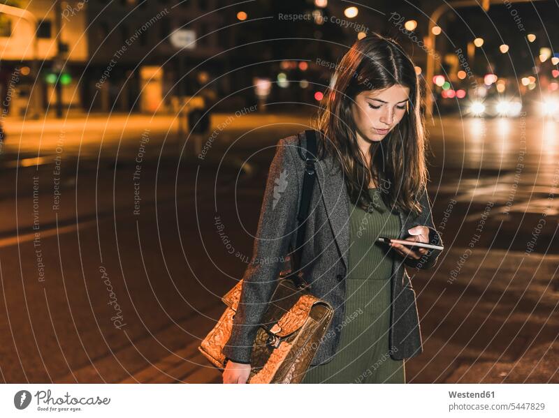 Junge Geschäftsfrau mit Ledertasche betrachtet Handy bei Nacht Frau weiblich Frauen nachts Erwachsener erwachsen Mensch Menschen Leute People Personen