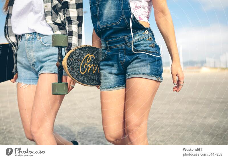 Zwei junge Frauen mit Longboards vor der Strandpromenade, Teilansicht Freundinnen Freunde Freundschaft Kameradschaft Hot Pants tragen transportieren