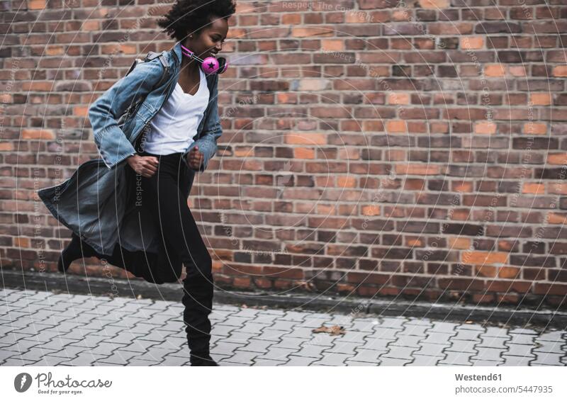 Lächelnde junge Frau mit Kopfhörern und Rucksack läuft auf dem Bürgersteig laufen rennen weiblich Frauen Erwachsener erwachsen Mensch Menschen Leute People