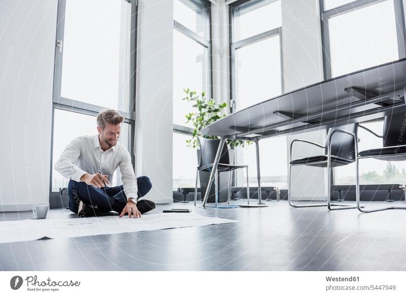 Lächelnder Geschäftsmann sitzt auf dem Boden seines Büros und betrachtet den Bauplan Businessmann Businessmänner Geschäftsmänner Geschäftsleute