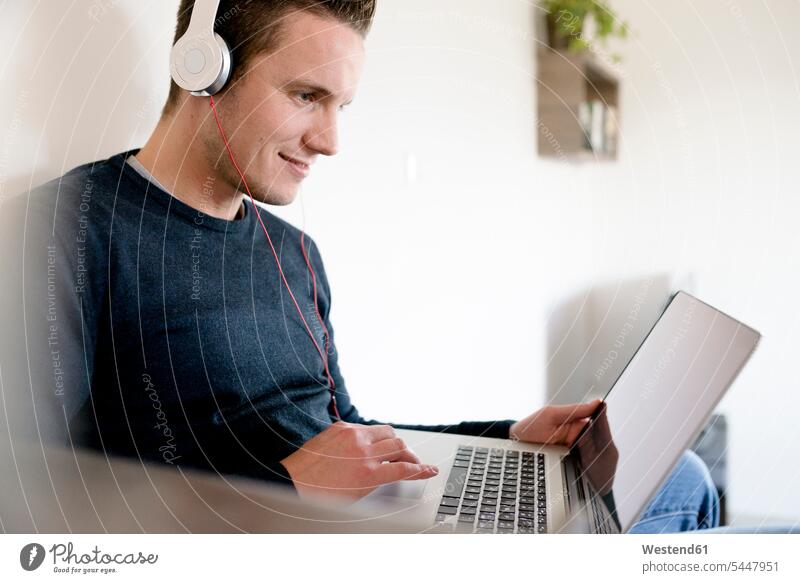 Mann benutzt Laptop und Kopfhörer zu Hause Männer männlich Wohnzimmer Wohnraum Wohnung Wohnen Wohnräume Wohnungen Kopfhoerer Notebook Laptops Notebooks