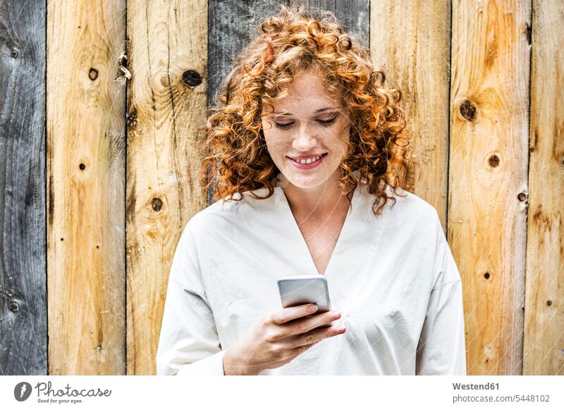 Porträt einer lächelnden jungen Frau, die vor einer Holzwand auf ein Handy schaut weiblich Frauen Smartphone iPhone Smartphones Portrait Porträts Portraits