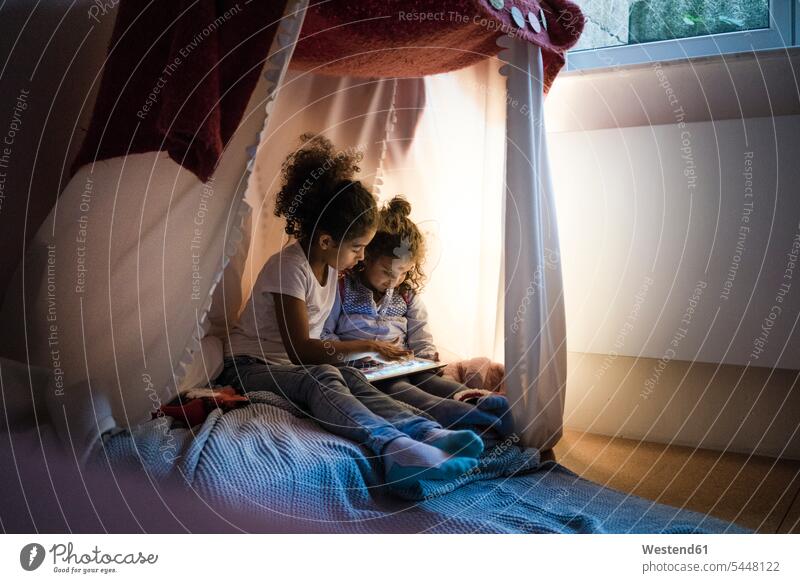 Zwei Schwestern sitzen im dunklen Kinderzimmer und schauen auf ein digitales Tablet sitzend sitzt spielen lesen Lektüre dunkel Geschwister Familie Familien