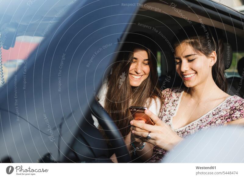 Zwei glückliche junge Frauen betrachten Handy im Auto lächeln Freundinnen Wagen PKWs Automobil Autos Mobiltelefon Handies Handys Mobiltelefone Freunde