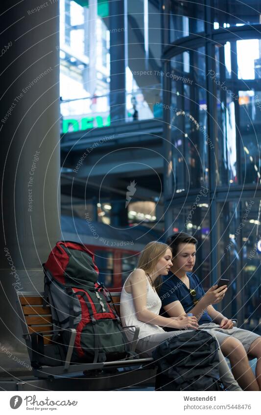 Junges Paar mit Rucksäcken sitzt mit Smartphone am Bahnhof Bahnhöfe Bahnhoefe besichtigen Besichtigung Reisende Reisender sitzen sitzend Rucksacktourist