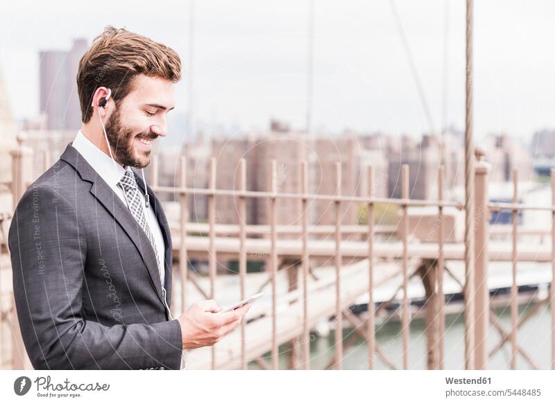 USA, New York City, lächelnder Geschäftsmann mit Handy und Ohrstöpseln auf der Brooklyn Bridge Brücke Bruecken Brücken Mobiltelefon Handies Handys Mobiltelefone