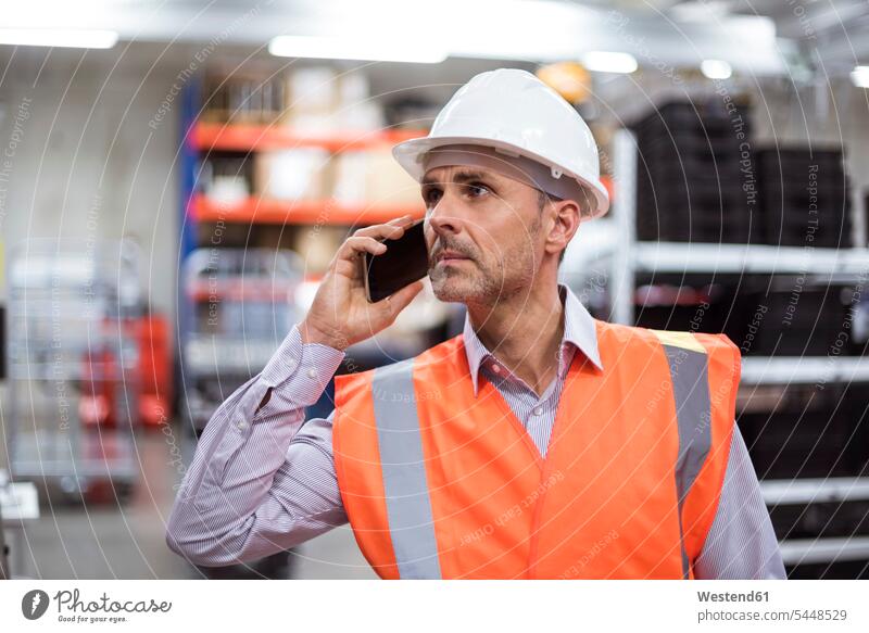 Mann in der Fabrikhalle mit Sicherheitsweste und Schutzhelm, der mit dem Handy telefoniert Männer männlich telefonieren anrufen Anruf telephonieren Mobiltelefon