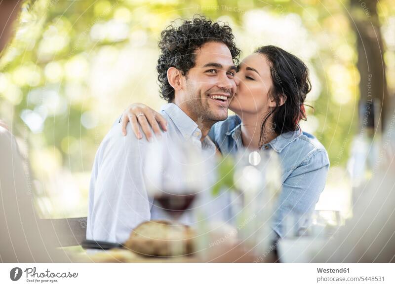 Junge Frau küsst einen glücklichen jungen Mann am Tisch im Freien Wein Weine lächeln küssen Küsse Kuss Rotwein Rotweine Paar Pärchen Paare Partnerschaft feiern