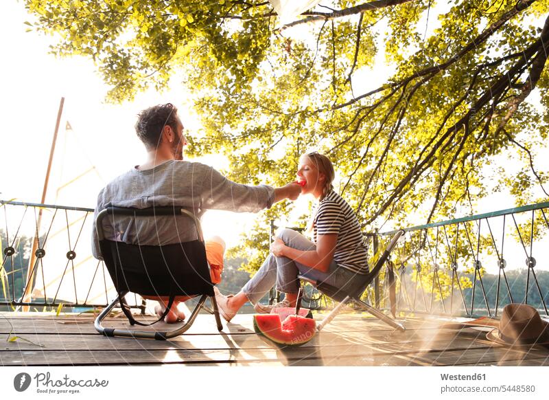 Junges Paar sitzt auf einem Steg an einem See und isst Wassermelone Stege Anlegestelle sitzen sitzend essen essend Citrullus lanatus Wassermelonen Pärchen Paare