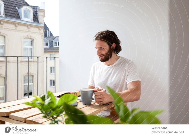 Lächelnder Mann sitzt mit Kaffeetasse auf dem Balkon und liest Zeitschrift Zeitschriften Magazin Magazine Männer männlich Balkone Erwachsener erwachsen Mensch