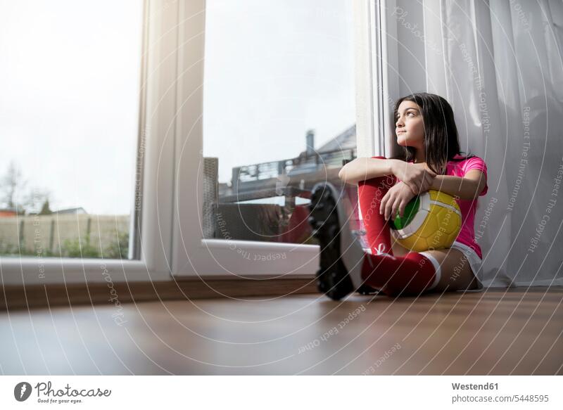 Mädchen in Fußballkleidung sitzt auf dem Boden im Wohnzimmer und schaut aus dem Fenster sitzen sitzend weiblich Fußbälle Fussball Kind Kinder Kids Mensch