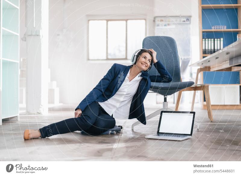 Lächelnde Geschäftsfrau, die auf dem Boden in einem Loft sitzt und mit Kopfhörern Musik hört Geschäftsfrauen Businesswomen Businessfrauen Businesswoman Laptop