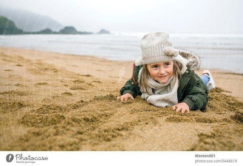 Glückliches Mädchen liegt im Winter am Strand im Sand sandig weiblich Beach Straende Strände Beaches glücklich glücklich sein glücklichsein liegen liegend Kind