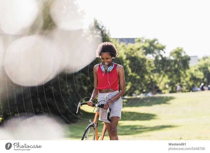 Lächelnde sportliche junge Frau schiebt Fahrrad im Park weiblich Frauen Bikes Fahrräder Räder Rad trainieren Parkanlagen Parks lächeln Erwachsener erwachsen