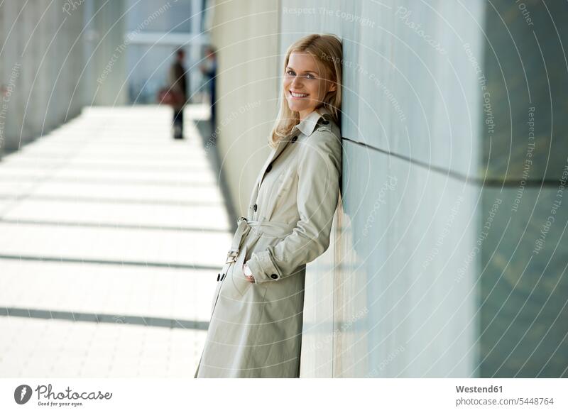 Porträt einer lächelnden blonden Frau in Trenchcoat, die sich an die Fassade lehnt Portrait Porträts Portraits weiblich Frauen Erwachsener erwachsen Mensch