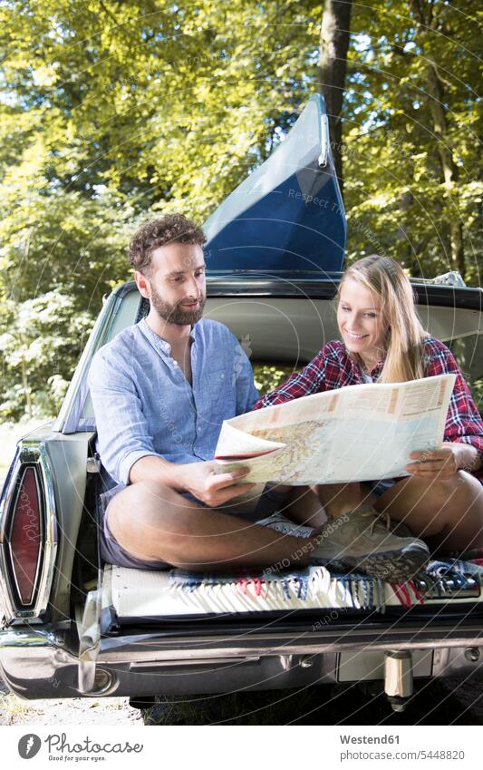 Lächelndes junges Paar mit Karte und Kanu im Auto an einem Bach Kanus lächeln Pärchen Paare Partnerschaft Wagen PKWs Automobil Autos Wald Forst Wälder Landkarte