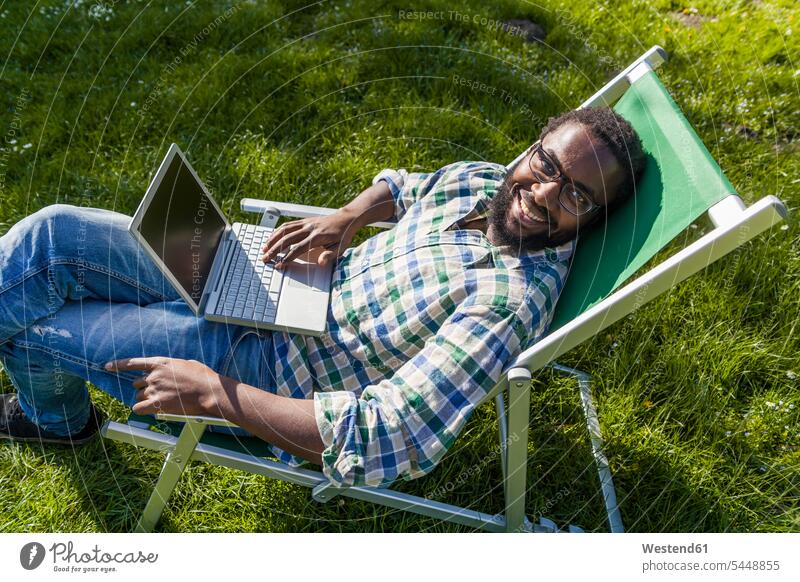 Lächelnder Mann mit Laptop sitzt auf einem Liegestuhl auf einer Wiese und schaut zur Kamera Männer männlich Notebook Laptops Notebooks Liegestühle Erwachsener