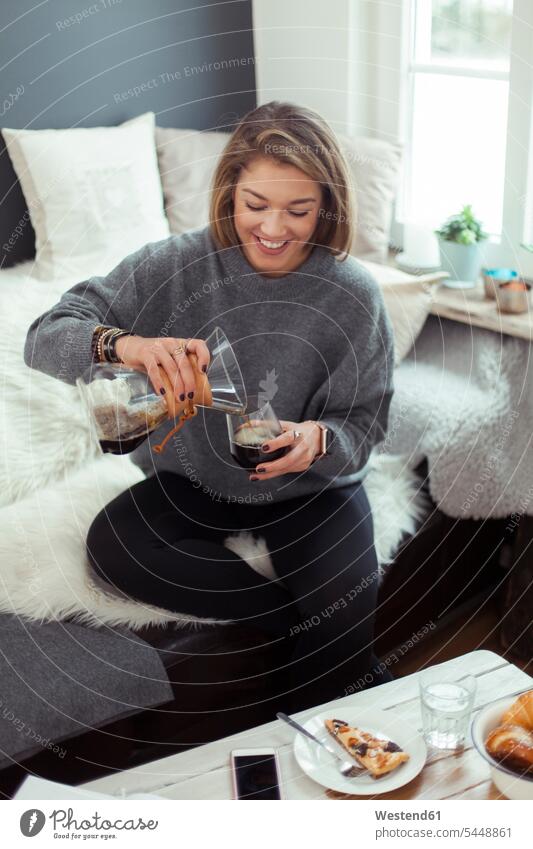 Lächelnde blonde Frau sitzt auf der Couch und gießt Kaffee in ein Glas weiblich Frauen Getränk Getraenk Getränke Getraenke Food and Drink Lebensmittel