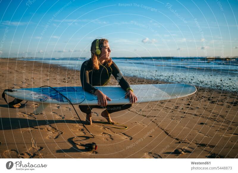 Junge Frau mit Surfbrett, die bei Sonnenuntergang am Strand Musik hört Surfbretter surfboard surfboards Surferin Wellenreiterinnen Surferinnen Kopfhörer