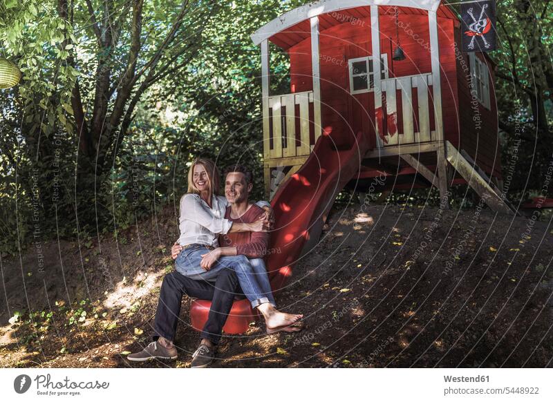 Glückliches Paar sitzt auf der Rutsche eines Gartenhäuschens im Wald sitzen sitzend Rutschen Gartenhaus Gartenhütte Pärchen Paare Partnerschaft lächeln Forst