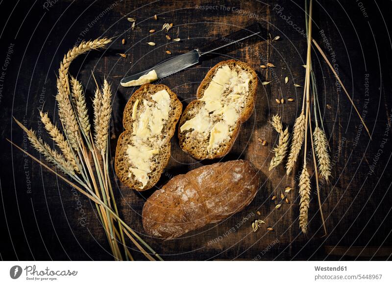 Gebuttertes Brot und Weizenähren auf dunklem Holz Draufsicht Vogelperspektive von oben Aufsicht Messer reif Reife Kulturpflanze Kulturpflanzen Getreide