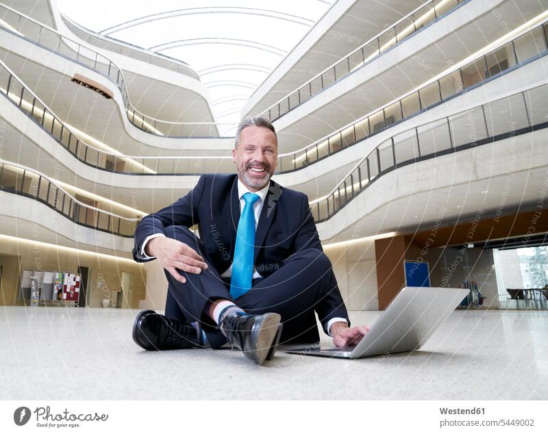 Glücklicher Geschäftsmann sitzt in modernem Bürogebäude mit Laptop auf dem Boden sitzen sitzend lächeln Notebook Laptops Notebooks Businessmann Businessmänner
