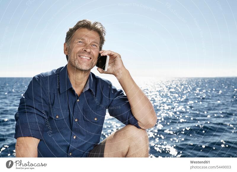 Porträt eines lächelnden reifen Mannes am Telefon vor dem Meer Männer männlich telefonieren anrufen Anruf telephonieren Portrait Porträts Portraits Erwachsener