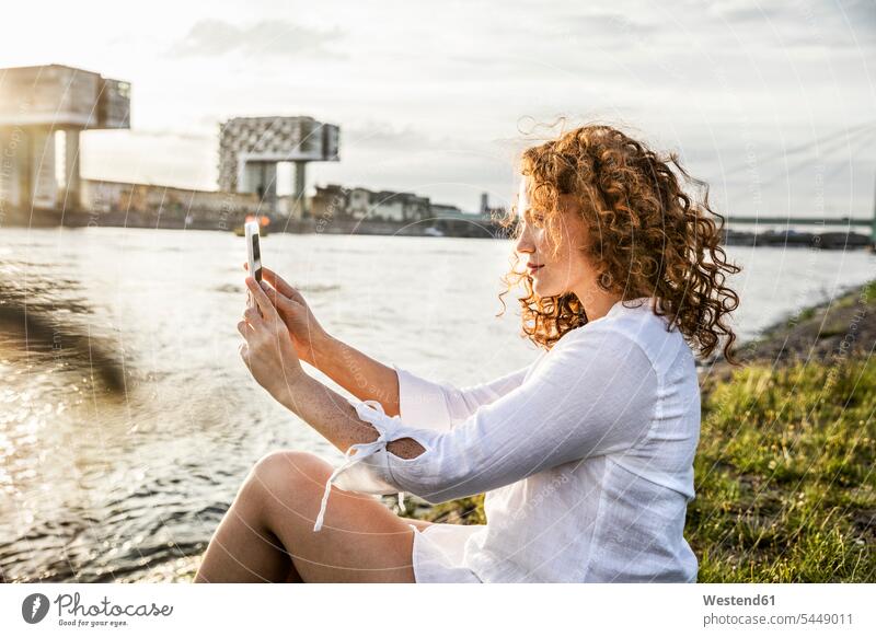 Deutschland, Köln, junge Frau sitzt abends am Flussufer und macht ein Selfie mit Handy weiblich Frauen Selfies Erwachsener erwachsen Mensch Menschen Leute
