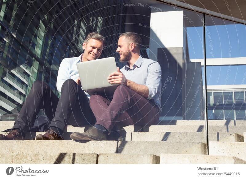 Zwei lächelnde Geschäftsleute sitzen mit Laptop auf der Treppe Notebook Laptops Notebooks Treppenaufgang sitzend sitzt Geschäftsmann Businessmann Businessmänner