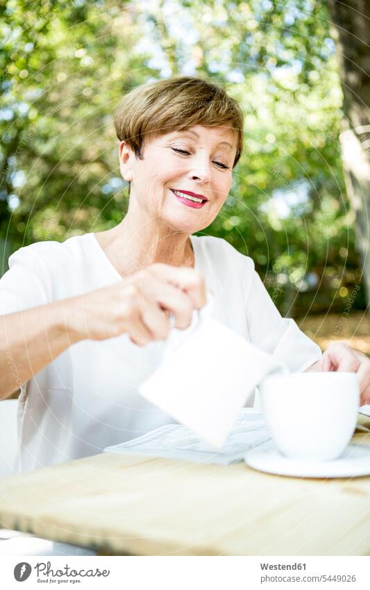 Lächelnde ältere Frau gießt im Freien Milch in eine Tasse Kaffee lächeln weiblich Frauen eingießen schütten einschenken schuetten Seniorin Seniorinnen alt
