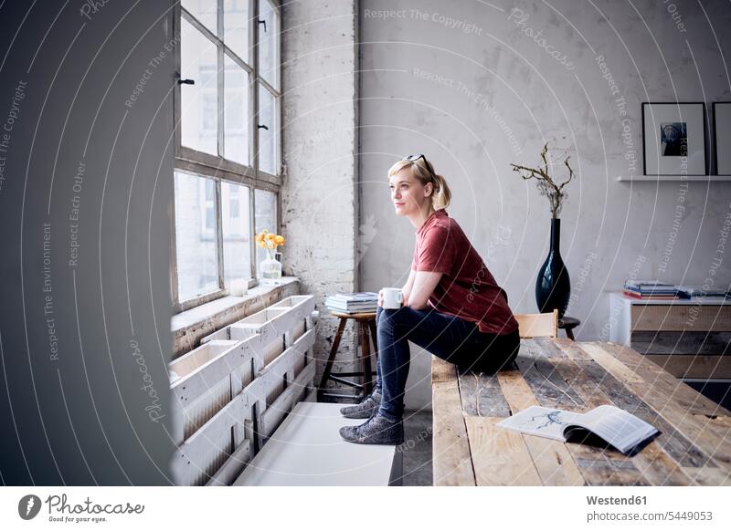 Lächelnde Frau mit Kaffeetasse sitzt auf dem Schreibtisch auf dem Dachboden und schaut durchs Fenster weiblich Frauen Loft Lofts Freiberufler freiberuflich