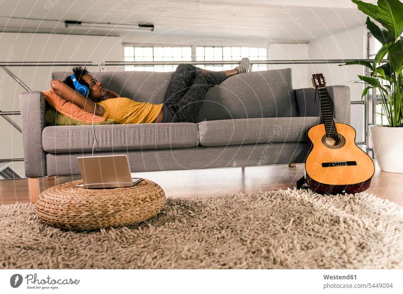 Junge Frau auf Couch liegend mit Kopfhörern an Laptop angeschlossen Sofa Couches Liege Sofas Kopfhoerer Notebook Laptops Notebooks liegt Gitarre Gitarren