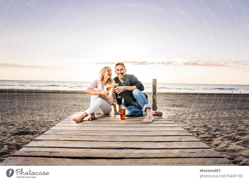 Glückliches Paar mit Weingläsern sitzt bei Sonnenuntergang auf der Strandpromenade Weine Pärchen Paare Partnerschaft sitzen sitzend Beach Straende Strände
