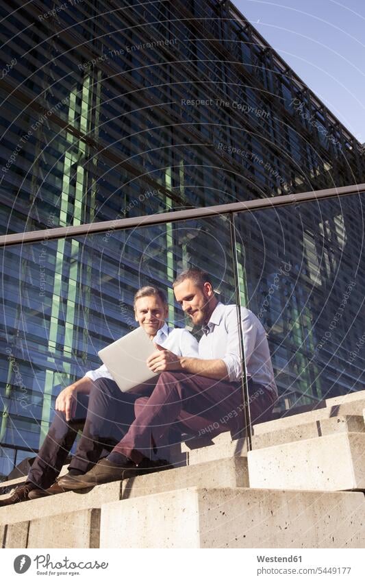 Zwei lächelnde Geschäftsleute sitzen mit Laptop auf der Treppe Kollegen Arbeitskollegen Geschäftsmann Businessmann Businessmänner Geschäftsmänner Notebook