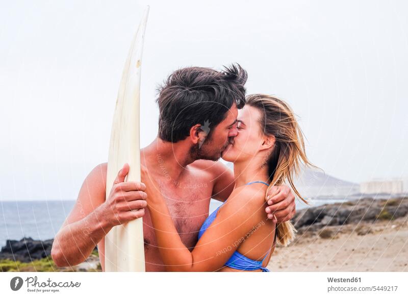 Junges Paar mit Surfbrett küsst sich am Strand Surfbretter surfboard surfboards küssen Küsse Kuss Liebe lieben Pärchen Paare Partnerschaft Surfen Surfing