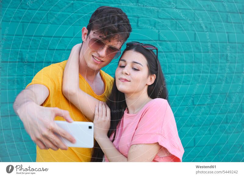 Junges Paar, das ein Selfie mit Smartphone vor einer blauen Backsteinmauer macht Pärchen Paare Partnerschaft Selfies iPhone Smartphones Mensch Menschen Leute