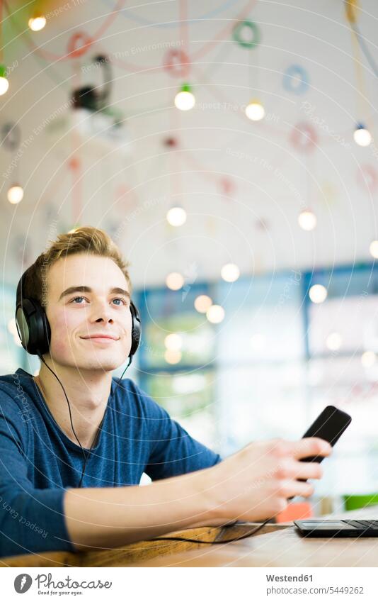 Porträt eines entspannten jungen Mannes, der in einem Café mit Kopfhörern und Smartphone Musik hört Kopfhoerer Portrait Porträts Portraits lächeln Männer