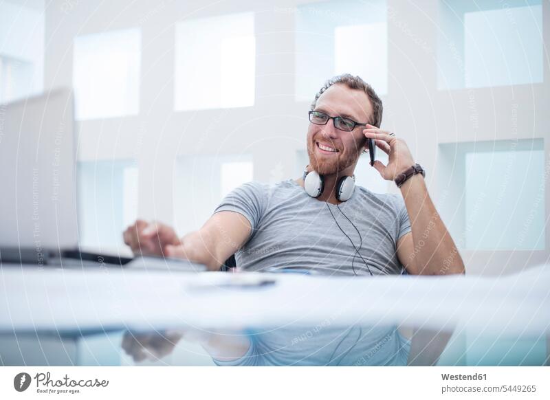 Junger Mann mit Kopfhörern um den Hals telefoniert cool Coolness telefonieren anrufen Anruf telephonieren Büro Office Büros arbeiten Arbeit Schreibtisch