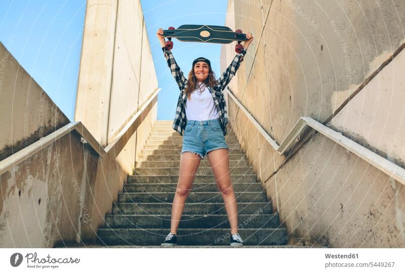 Porträt einer glücklichen jungen Frau, die auf einer Treppe steht und ihr Longboard hochhält Skateboarderin Skateboardfahrerin Skaterin Skateboarderinnen