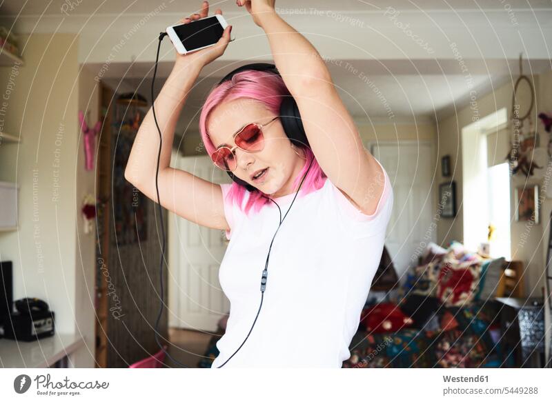 Enthusiastische junge Frau mit rosa Haaren, die zu Hause Musik hört Brille Brillen Kopfhörer Kopfhoerer weiblich Frauen hören hoeren Handy Mobiltelefon Handies