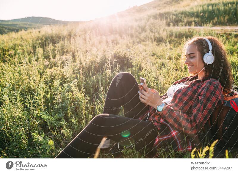 Teenagerin, die auf einer Wiese mit Kopfhörern Musik hört und auf ihr Handy schaut junges Mädchen Teenagerinnen weiblich junge Frau Kopfhoerer Jugendliche