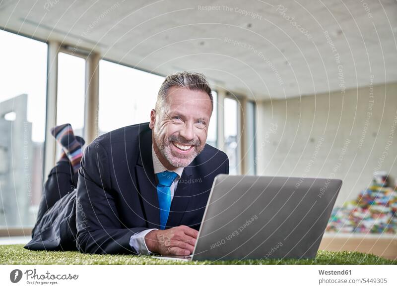 Glücklicher Geschäftsmann liegt mit Laptop auf Kunstrasen Businessmann Businessmänner Geschäftsmänner Notebook Laptops Notebooks lächeln Geschäftsleute