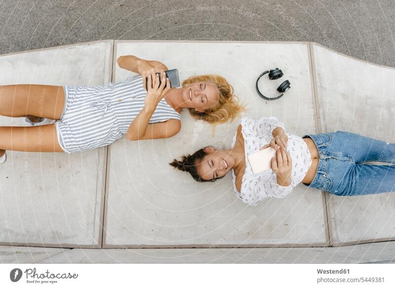 Zwei glückliche junge Frauen mit Mobiltelefonen liegen auf einer Rampe in einem Skatepark Freundinnen weiblich Handy Handies Handys Auffahrt Skateboardpark