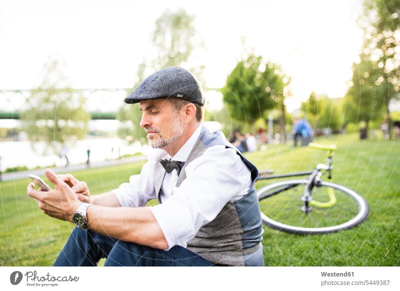 Selbstbewusster reifer Geschäftsmann mit Fahrrad und Smartphone im Stadtpark auf dem Rasen sitzend Handy Mobiltelefon Handies Handys Mobiltelefone Businessmann