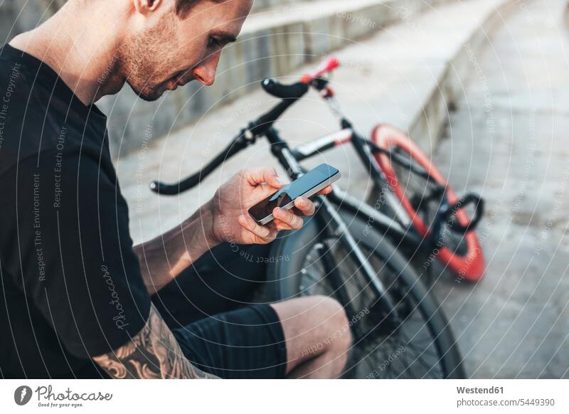 Junger Mann sitzt neben einem Fixie-Bike und benutzt sein Handy sitzen sitzend Männer männlich Fahrrad Bikes Fahrräder Räder Rad Mobiltelefon Handies Handys