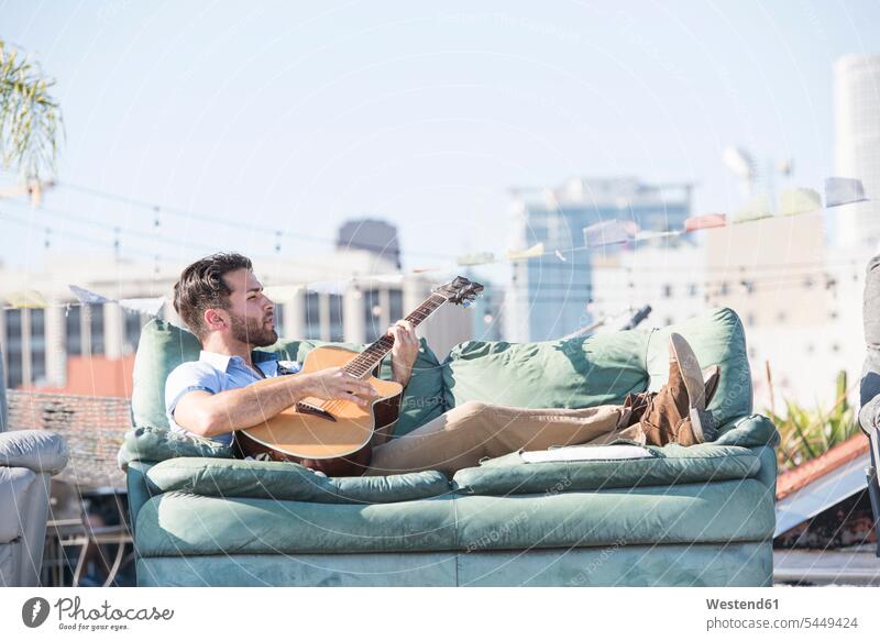 Junger Mann auf dem Dach, der auf dem Sofa liegt und Gitarre spielt Couches Liege Sofas Musiker liegen liegend Dachterrasse Dachterrassen spielen Gitarren