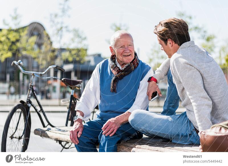 Älterer Mann und erwachsener Enkel unterhalten sich auf einer Bank sitzen sitzend sitzt lächeln glücklich Glück glücklich sein glücklichsein Enkelsöhne