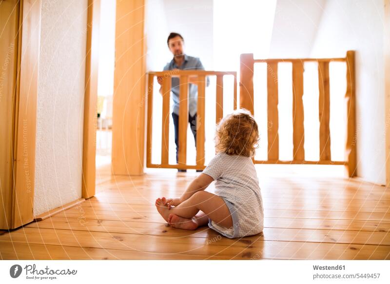 Kleiner Junge sitzt auf Holzboden und schaut den Vater hinter der Absperrung an der Treppe an Baby Babies Babys Säuglinge Kind Kinder Treppenaufgang Barriere
