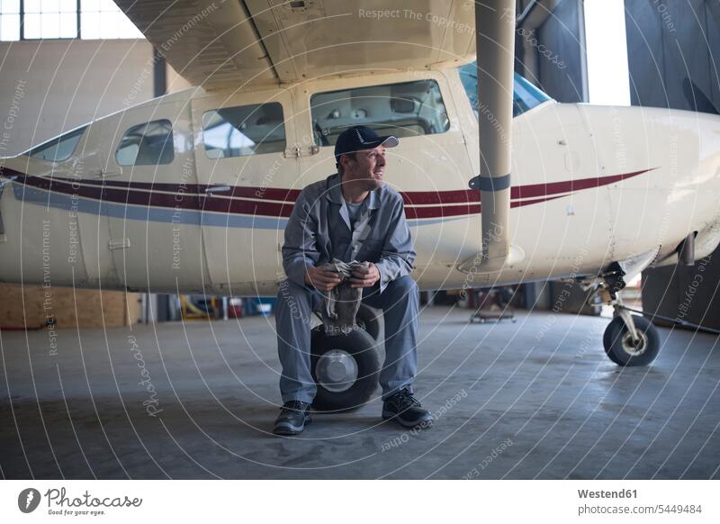 Mechaniker sitzt neben einem Leichtflugzeug im Hangar des Flugplatzes Flugzeug Flieger Flugzeuge Monteur Hangars Flugzeughalle Flugzeughallen sitzen sitzend