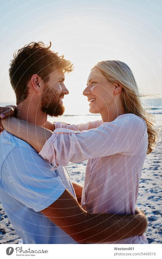 Lächelndes Paar umarmt sich am Strand bei Sonnenuntergang lächeln umarmen Umarmung Umarmungen Arm umlegen Beach Straende Strände Beaches glücklich Glück